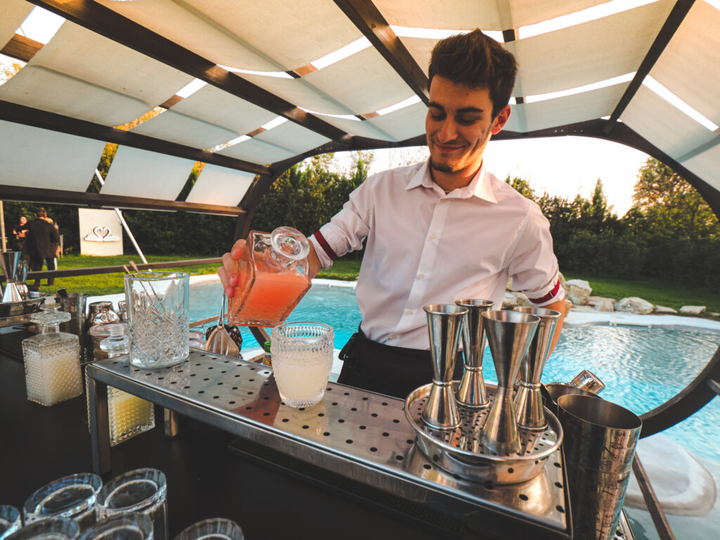 A bartender skillfully mixing drinks at an outdoor bar near a pool, with the sun setting in the background.